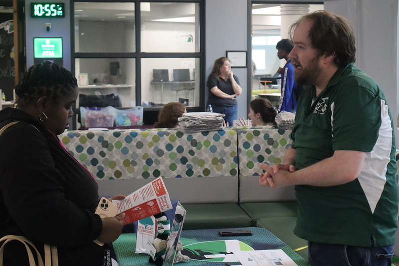 Two people standing at a table having a discussion. The person on the left is reading over a pamphlet while the person on the right is speaking