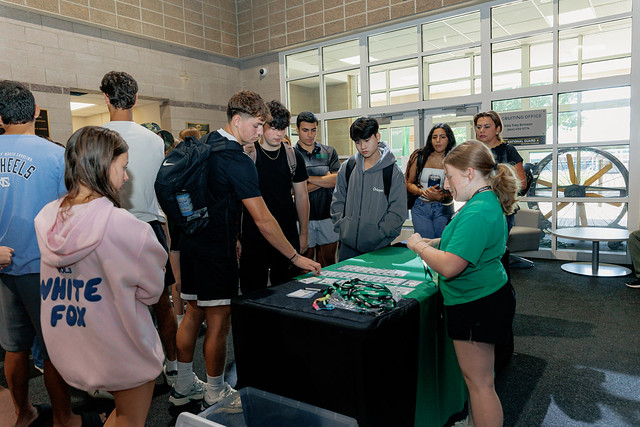 A group of incoming students standing around a registration table. An Upstate employee is helping them look at paperwork. One student is pointing to something on the page.