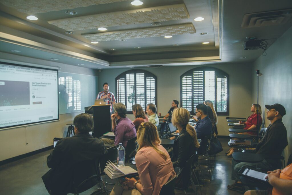 Faculty member speaking at a small podium to the side of projection slide. Three rows of students face the projection screen.