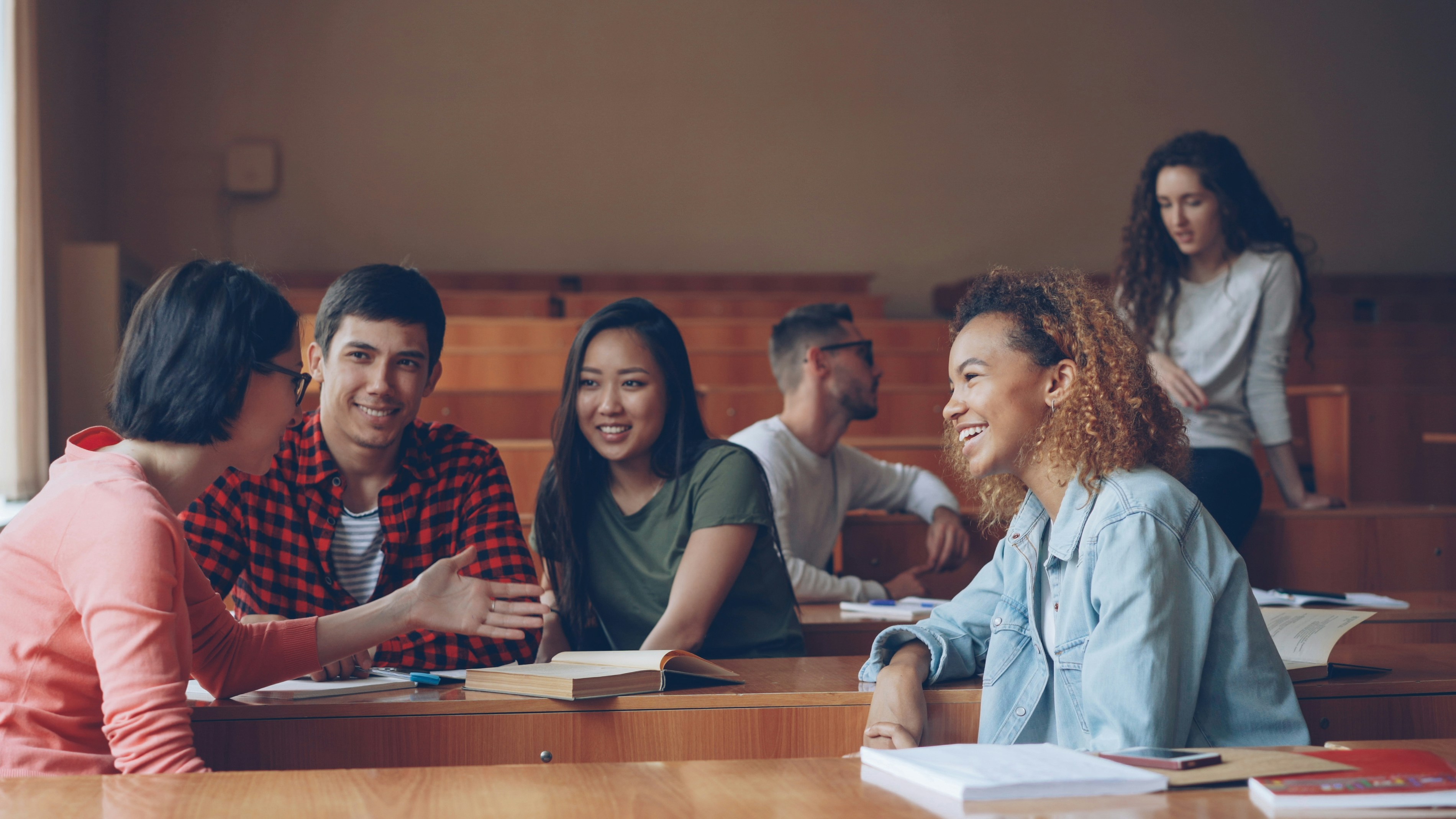 Several students are talking together in a lecture hall. There are books and notebooks on the table.